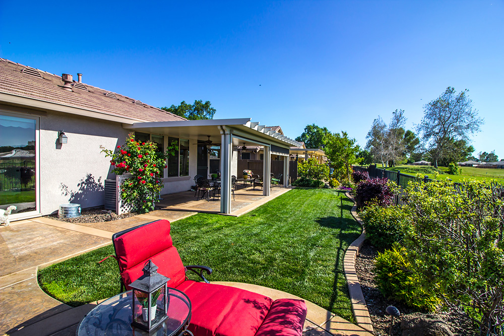 A serene backyard with a concrete patio, covered porch, manicured lawn, vibrant flower bushes, and a view of a golf course.