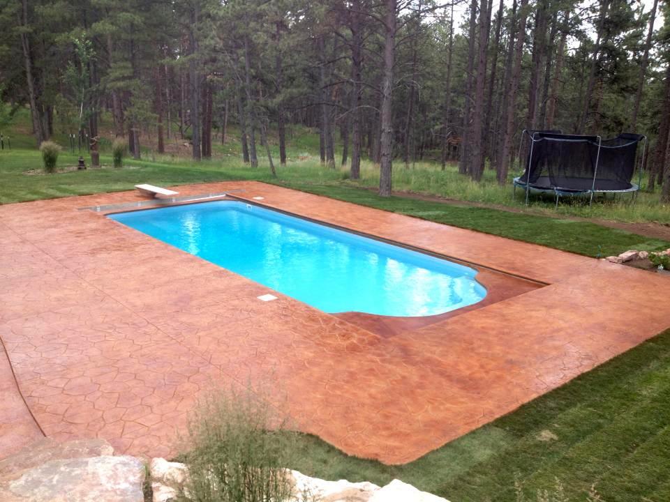 A rectangular swimming pool with a rust-colored stamped concrete deck in a forested backyard with a trampoline in the distance.