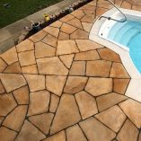 The image displays a decorative concrete pool deck with a flagstone pattern. The stones are in various shades of tan and beige, arranged in an irregular, interlocking design, enhancing the poolside area with a natural stone appearance.