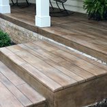 "Elegant outdoor steps with wood-patterned concrete design, bordered by a stone wall, leading to a columned porch.