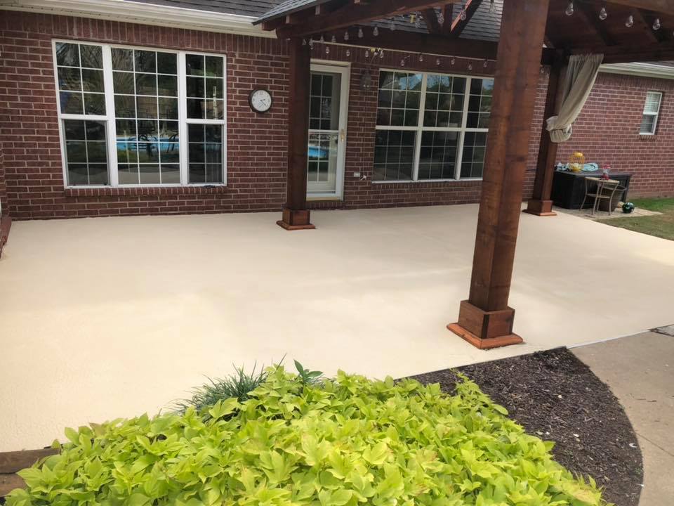 A freshly finished light-colored concrete patio with a pergola, adjacent to a brick house with white windows, overlooking a garden.