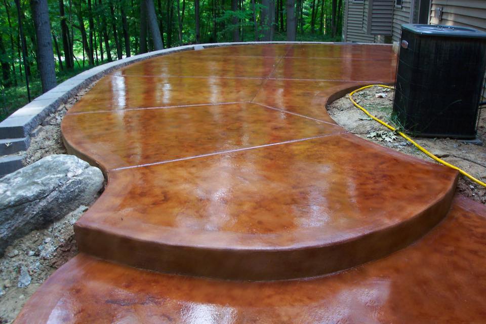 A curved, glossy stained concrete patio with a brown tone adjacent to a house with wooded surroundings.