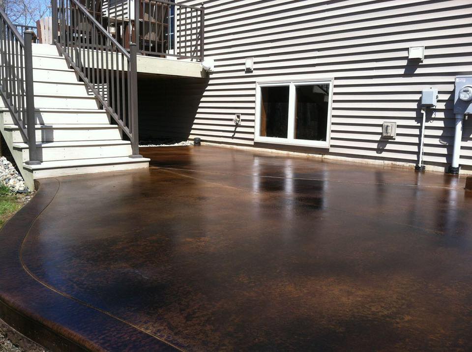 A shiny, stained concrete patio with curved edges, adjacent to a house with vinyl siding and wooden stairs.