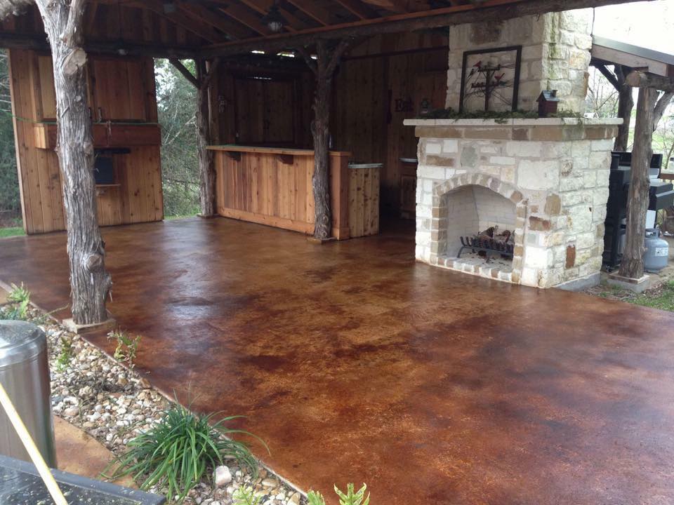 An outdoor covered patio area with a warm-toned stained concrete floor, featuring a stone fireplace and rustic wooden accents.