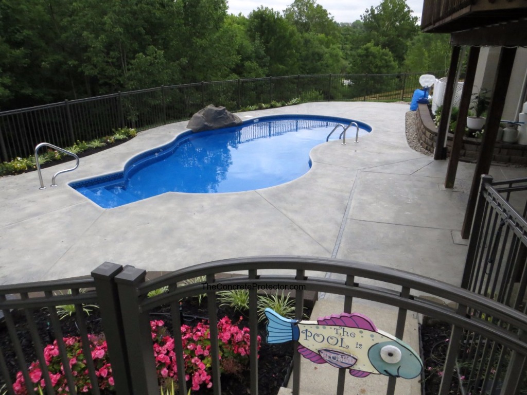 A kidney-shaped swimming pool with blue waters, surrounded by a grey concrete deck and metal fence, with lush greenery in the background.