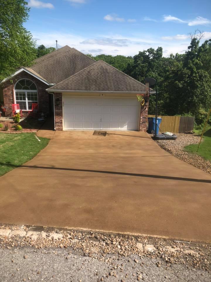 Brick house with a double garage door and a tan concrete driveway, surrounded by greenery and a clear blue sky.