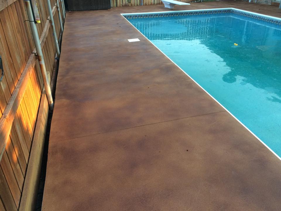 A backyard pool with a reddish-brown stamped concrete deck, wooden fence, and clear blue water reflecting the light.