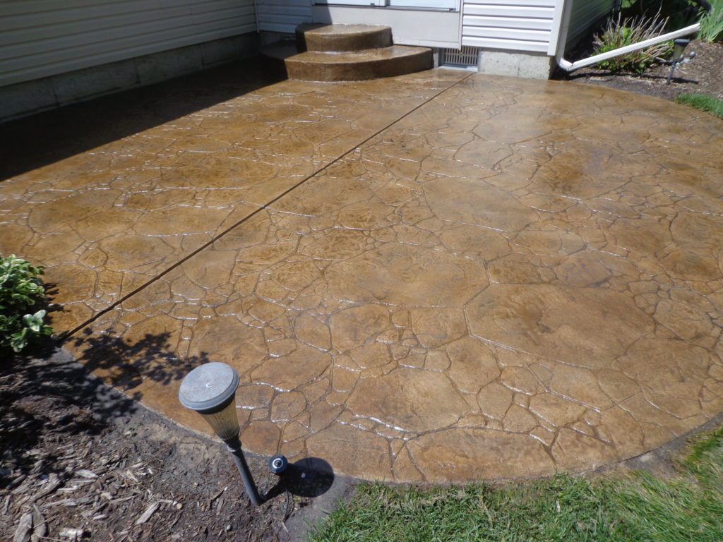 A golden-brown stamped concrete patio with a random stone pattern beside a house, with a solar path light and landscaping.