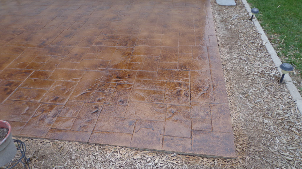 A wet stamped concrete surface with a rustic, brown coloration next to a landscaped area with mulch.