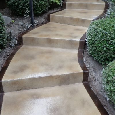 Textured concrete steps with a dual-tone border design, flanked by lush green shrubs and a metal handrail.