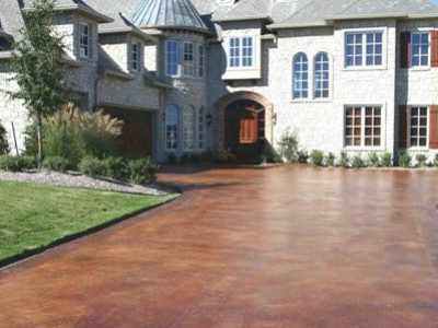 A grand stone house with large windows and a wooden door, featuring a spacious, russet-colored smooth concrete driveway.