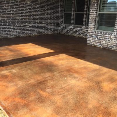 A shaded concrete patio with a rich, amber-colored stain, set against a dark brick home with contrasting white windows.